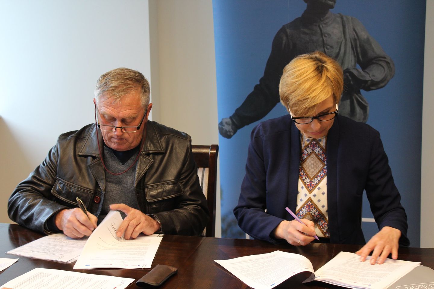 Na zdjęciu znajdują się dwie osoby podpisujące dokumenty.
