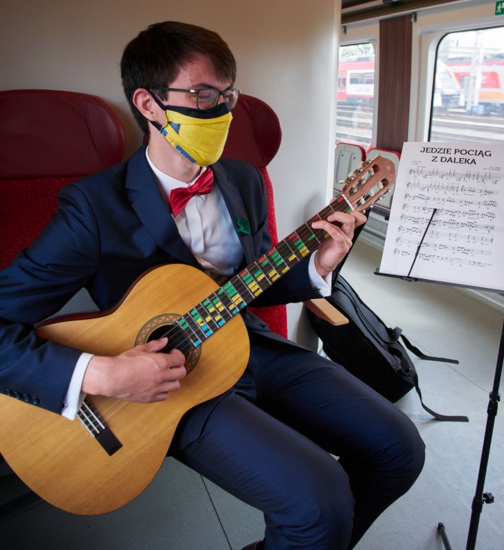 Na zdjęciu widać chłopaka siedzącego w pociągu i grającego na gitarze.